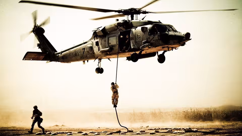Soldier rappelling from a hovering helicopter as another runs across dusty ground
