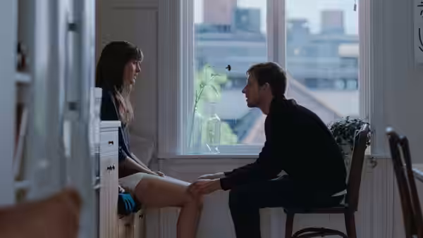 A man sits beside a woman, gently holding her leg in an apartment