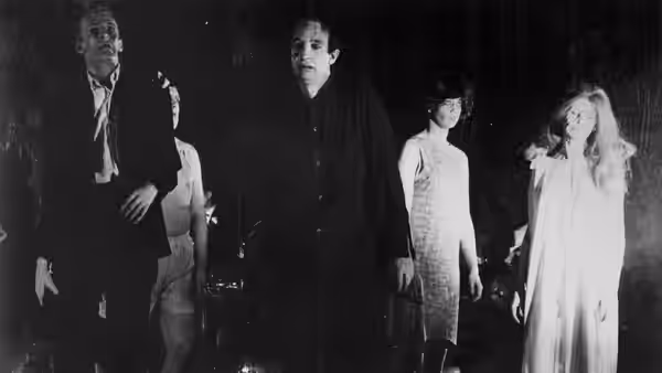 Several people stand in a dark room, including a woman in a white dress