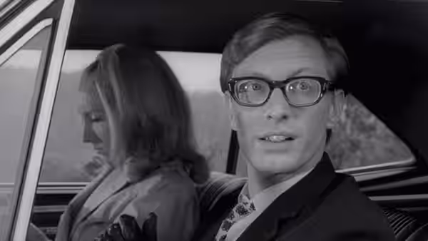 A bespectacled man sits beside a woman in the back of a car