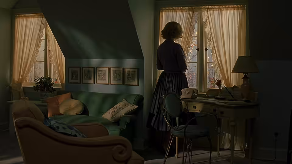 A woman in a blue dress stands by the window in a living room
