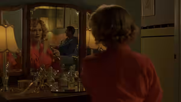 A woman in a red blouse looks at her reflection in a vanity mirror