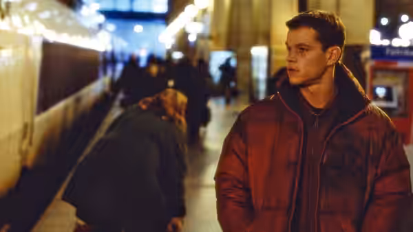 Jason Bourne standing on a train platform as a woman bends over