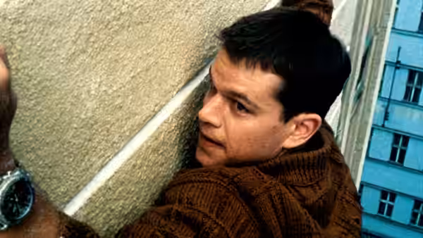 Jason Bourne peering around the corner of a concrete wall outdoors