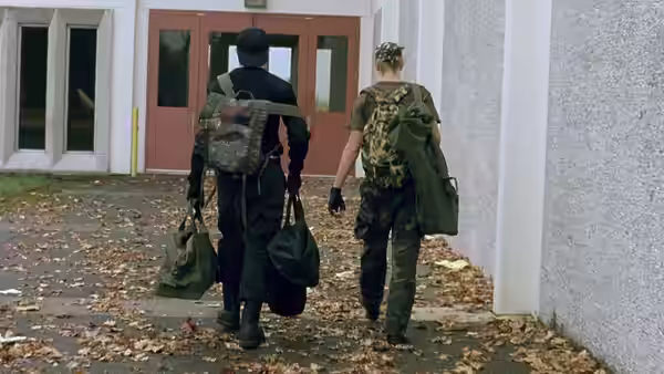 Two young men in camouflage clothing carrying bags walking towards a building