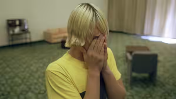 A young man with blonde hair covering his face with his hands while seated