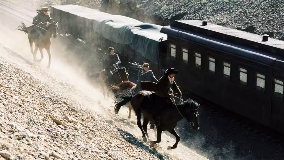 Mounted riders race alongside a passenger train on a dusty hillside