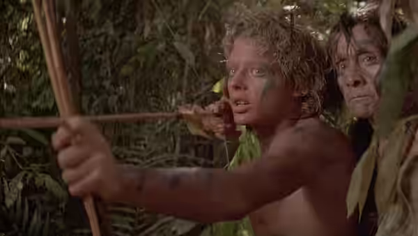 A young man aims a bow through the jungle while an older man watches