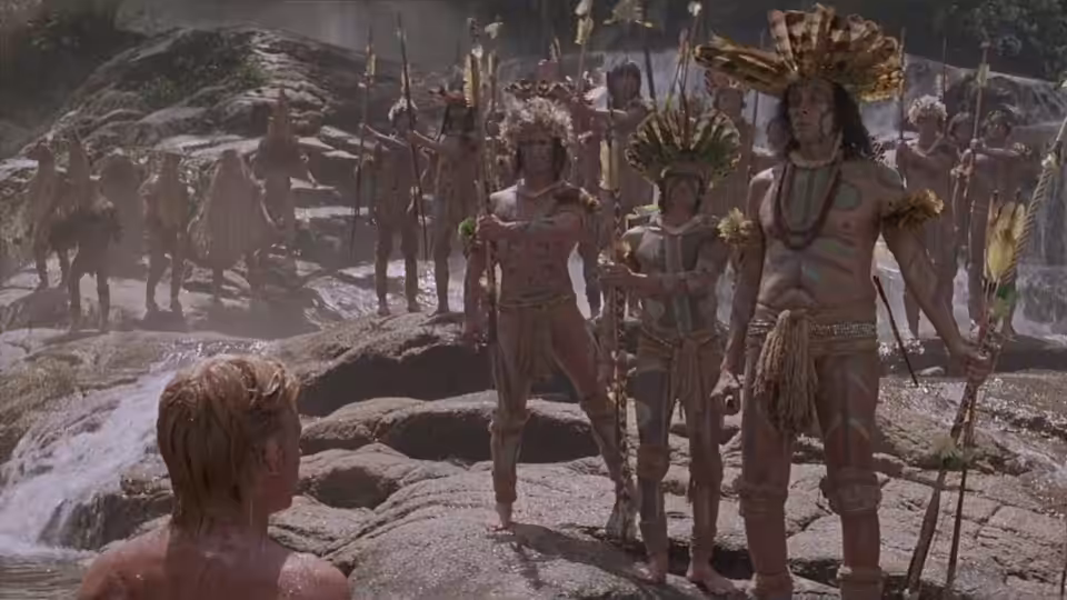 A blond man faces Indigenous people gathered among rocky riverbanks