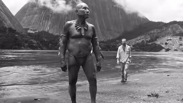 Indigenous man with adornments stands on riverbank as another walks by