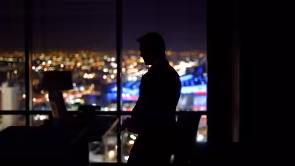 A man in a suit stands silhouetted before a window overlooking city lights at night