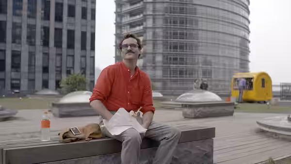 Theodore (Joaquin Phoenix) sits on a bench in an urban plaza holding papers