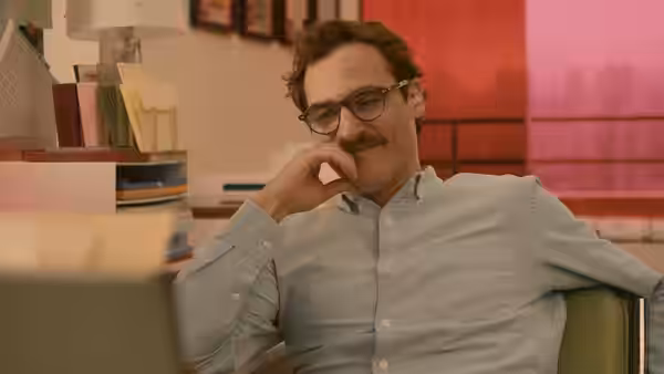 Theodore (Joaquin Phoenix) sits in an office chair smiling with hand near his chin
