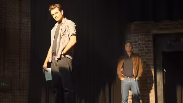 A young man standing on stage holding papers while another watches from the side