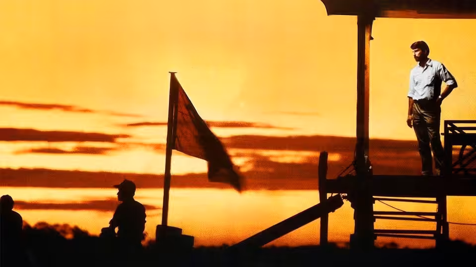 Man stands on wooden platform beside red flag at sunset with silhouetted figures below