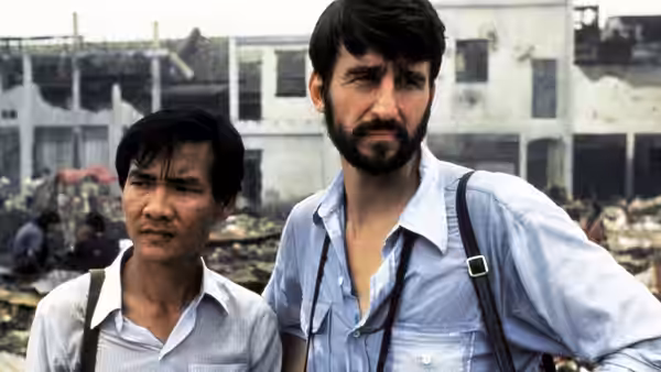 Bearded man and Asian companion stand amid rubble before destroyed buildings