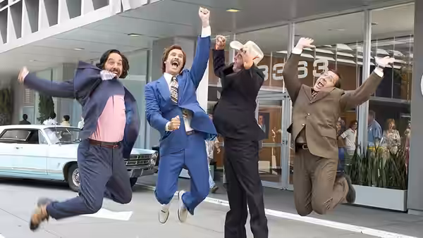 Ron Burgundy (Will Ferrell) and colleagues jumping with arms raised outside a building