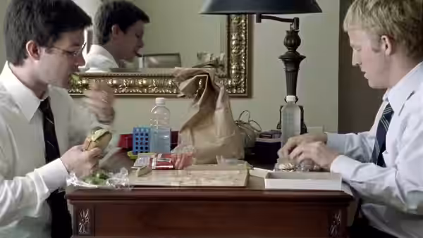 Aaron (Shane Carruth) handling small objects while another man eats at a table