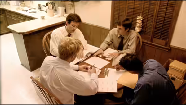 Four men reviewing documents spread across a kitchen table