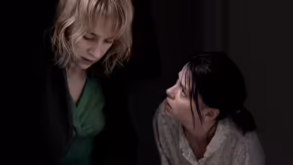 Blonde woman leans over dark-haired woman sitting on floor in dark room