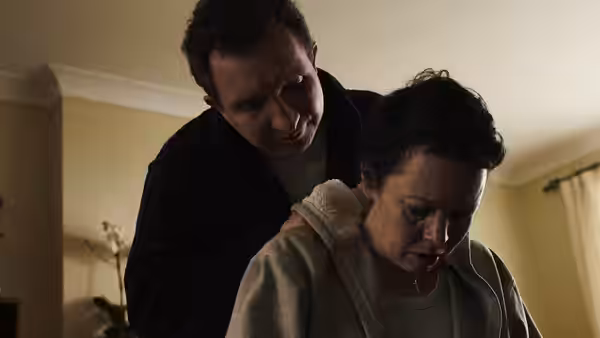 Hannah (Olivia Colman) with a man standing close behind her indoors