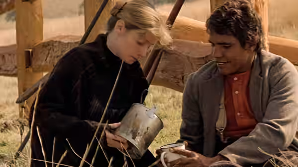 A blonde woman and a man sit beside a wooden fence, examining a metal container