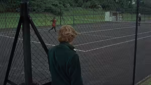 Thomas watches a mime play imaginary tennis behind a chain-link fence