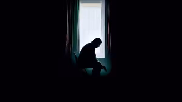 A man sits hunched in silhouette beside a curtained window in darkness