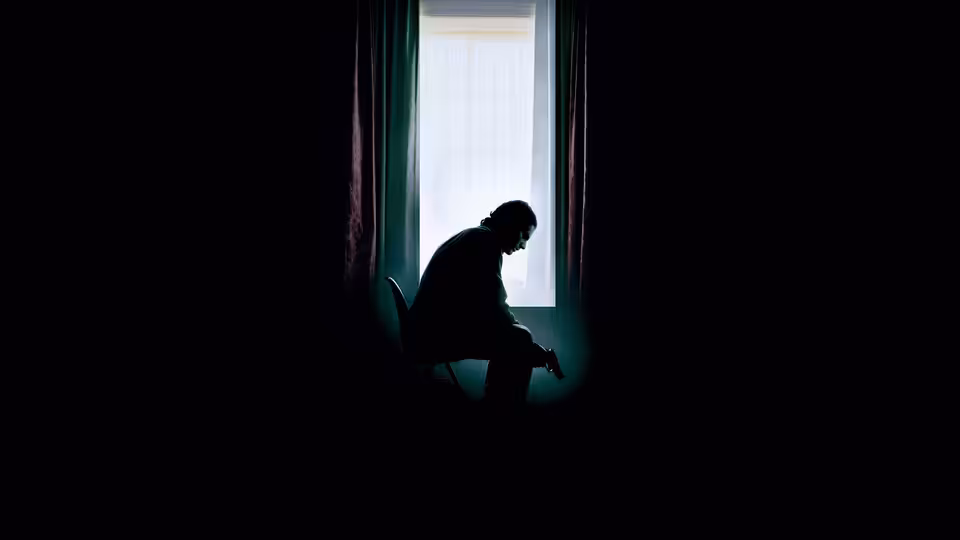 A man sits hunched in silhouette beside a curtained window in darkness