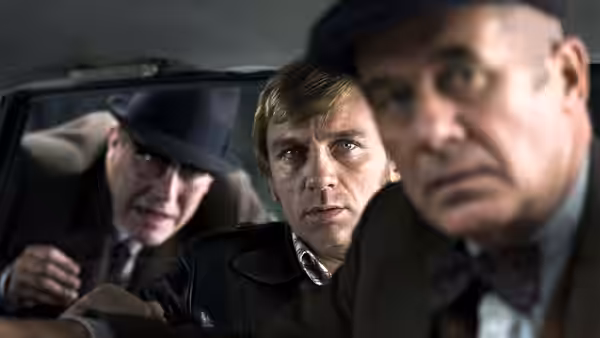 Three men sit inside a car looking forward with serious expressions
