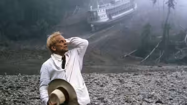 Fitzcarraldo (Klaus Kinski) stands on a rocky riverbank holding his hat