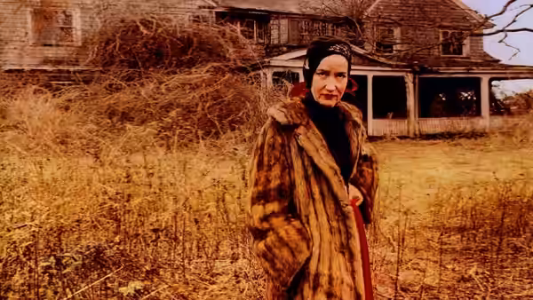 Woman in fur coat and headscarf stands in overgrown yard before dilapidated house