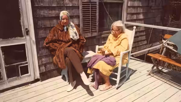 Two women sit on a wooden porch outside a house
