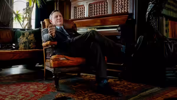 Benoit Blanc reclining in a chair and pointing in the study