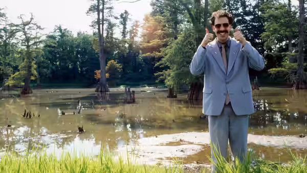 Borat (Sacha Baron Cohen) in a suit giving thumbs up in a swamp