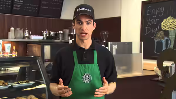 Nathan Fielder in green Starbucks apron gesturing with hands in a cafe