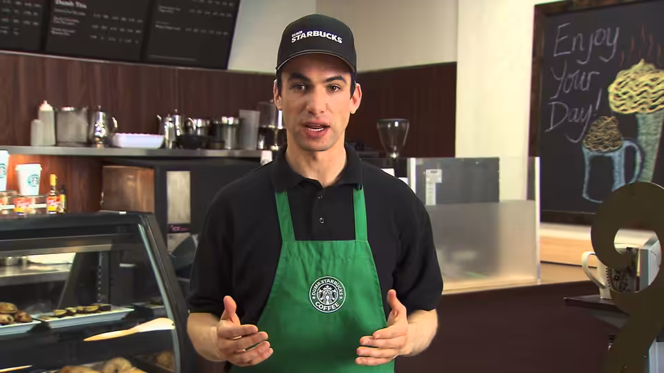 Nathan Fielder in green Starbucks apron gesturing with hands in a cafe