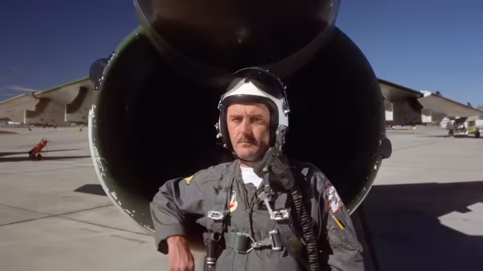 a pilot in flight suit standing before a large jet engine on tarmac