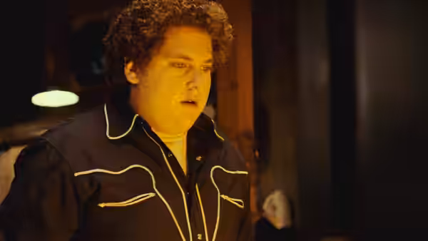 Seth (Jonah Hill) in a dark jacket standing indoors at night