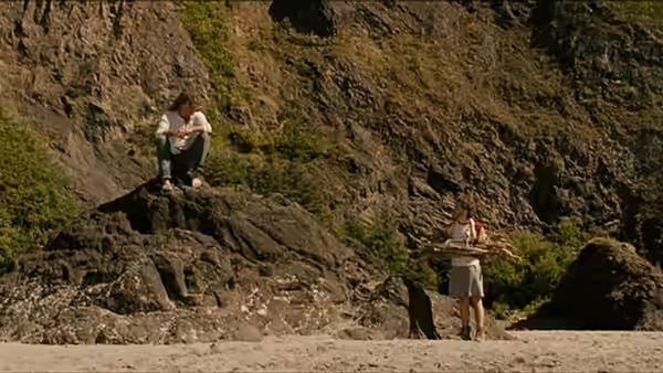 a man carrying firewood while a woman sits on rocks above the shore