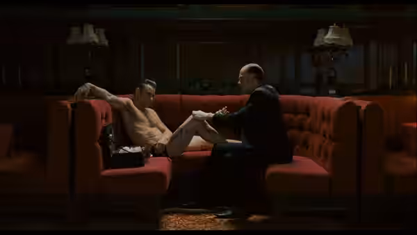 Shirtless man reclines on a red couch while another tends his foot