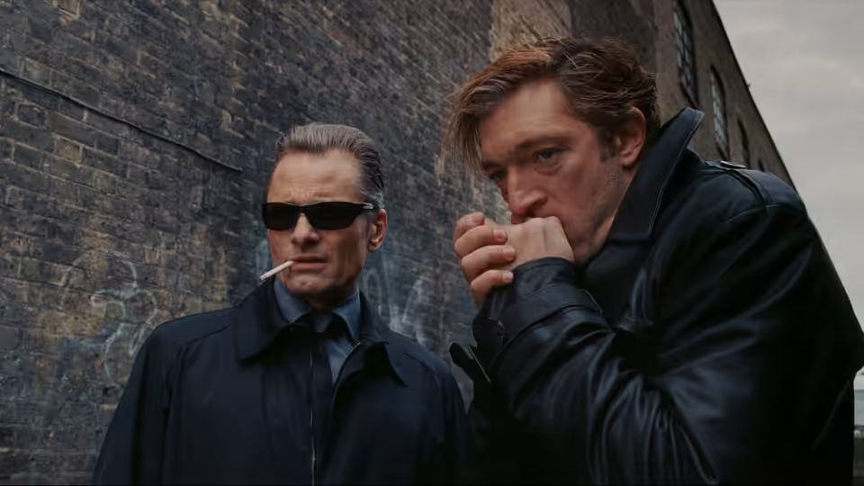 Kirill (Vincent Cassel) stands beside a man against a brick wall