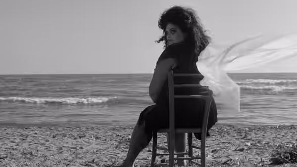 A dark-haired woman sits backward on a chair at the beach as sheer fabric blows behind her