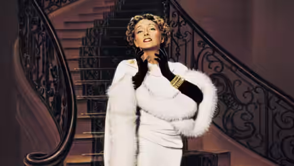 Norma Desmond (Gloria Swanson) poses in a white fur coat on her grand staircase
