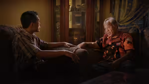 Two men sit on a sofa holding hands in a living room
