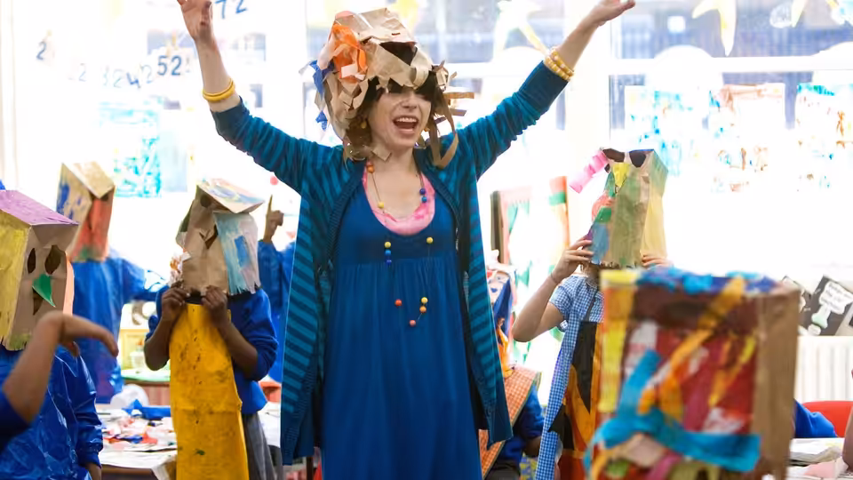 Poppy (Sally Hawkins) raising her arms joyfully among children wearing box hats in a classroom