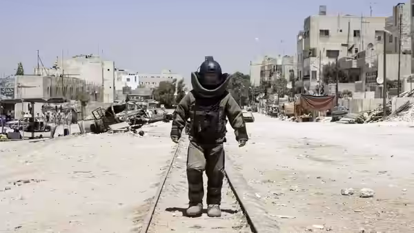 A soldier in a bomb suit stands on railway tracks amid ruined buildings