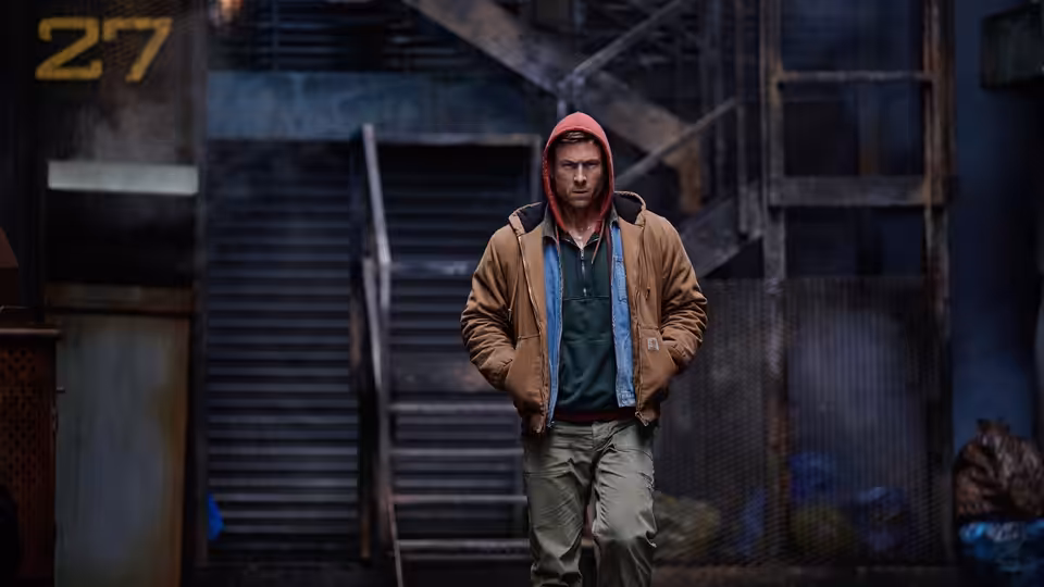 Ben Richards (Glen Powell) standing with hands in pockets before metal stairs