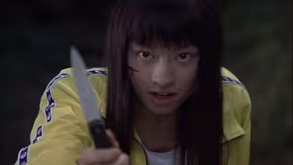 Young woman in yellow jacket holds knife forward with blood on face
