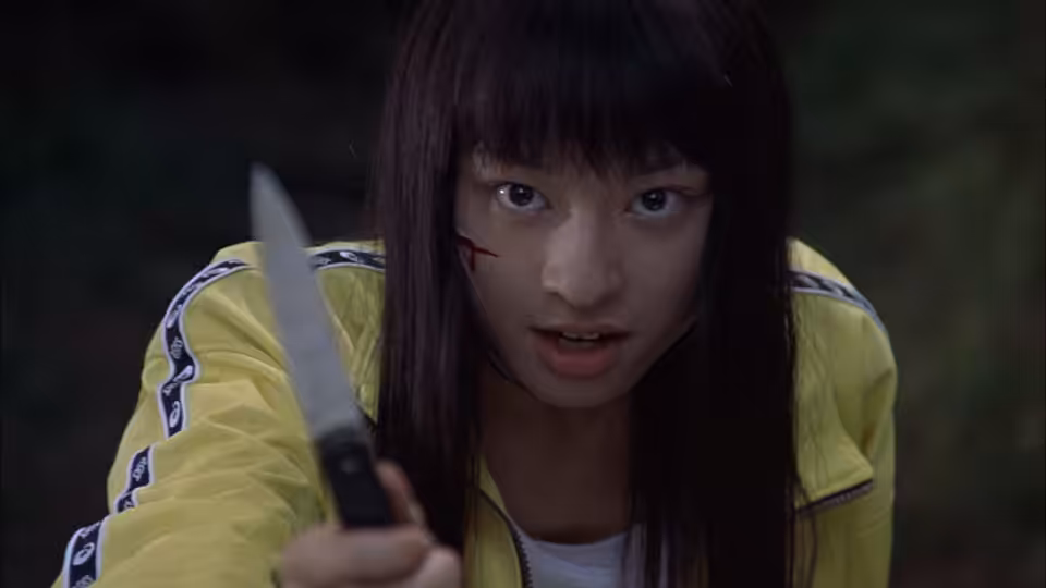 Young woman in yellow jacket holds knife forward with blood on face